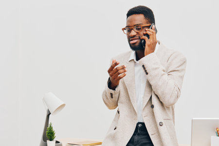 Confident smiling man with glasses in beige blazer talking on mobile phone while standing at workplace with modern desk and minimalistic decor. Business communication, professional lifestyle concept.の写真素材