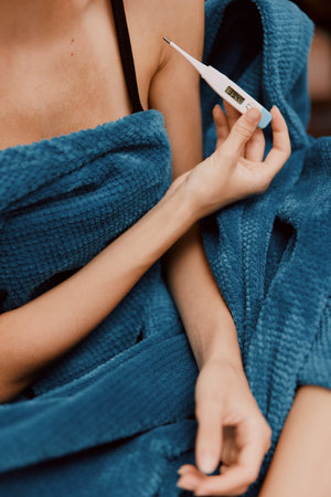 Woman wrapped in blue blanket holding a thermometer with another one in her earlobeの写真素材