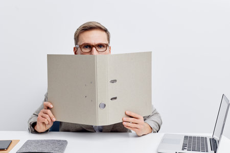 Young caucasian man working in cyberspace hold folder cover face glasses wear gray shirt sit at...