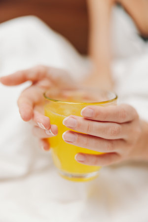 Woman holding glass of orange juice in bed with white sheetの写真素材