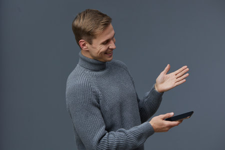 smiling man using smartphone hold in hand wearing grey knitted sweater dark background digital...