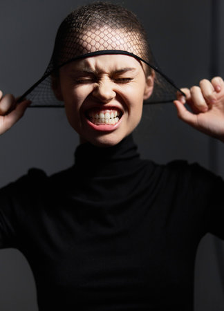 Emotional young woman in dark outfit expressing frustration while pulling on a mesh head covering....