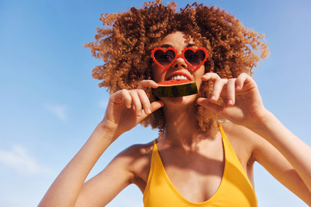 Young woman with curly hair wearing heart shaped sunglasses enjoys a slice of watermelon against a bright blue skyの写真素材