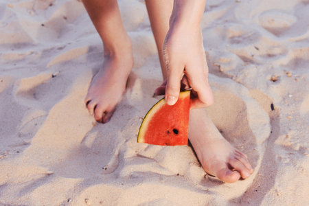 A person holding a slice of watermelon while standing on sandy beach The warm tones of the sand complement the vibrant color of the fruitの写真素材