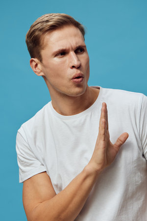 Thoughtful young man with short brown hair, wearing casual white t-shirt, expressing disapproval or hesitation, positioned against plain blue background, studio portrait. People emotion conceptの写真素材
