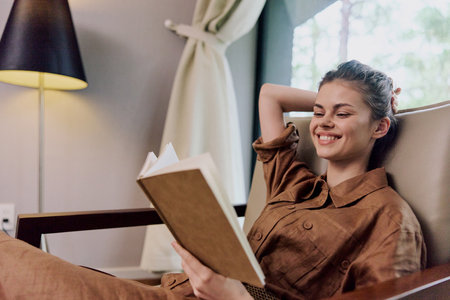 Relaxed young woman reading a book indoors, wearing casual brown outfit, with a warm smile, natural light and soft decor, perfect for promoting leisure and readingの写真素材