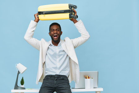 Cheerful young African American man in business casual attire holding a yellow suitcase overhead, standing in a modern office with blue background, expressive and joyful mood, concept of travel, success, or new beginnings.の写真素材