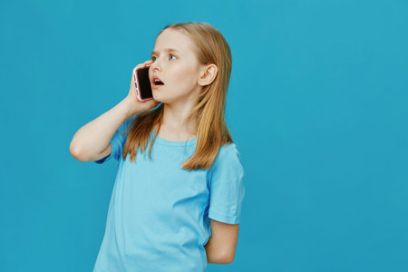 A young girl with light brown hair in a blue t shirt talking on a smartphone, expressing surprise against a bright blue background, ideal for concepts of communication and youthの写真素材