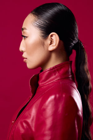 Fashionable asian woman in a red leather jacket and ponytail hairstyle looking off to the sideの写真素材