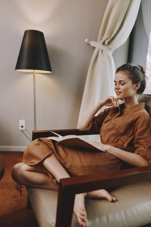 Relaxed young woman reading a book in a cozy room, wearing a brown dress, enjoying a peaceful moment...