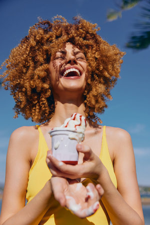 Joyful woman with curly hair holding an ice cream cone in sunlight outdoors