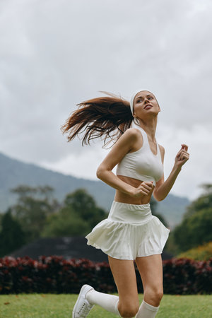 Active young woman running outdoors in sporty white outfit, expressing joy against a green nature background with mountains, promoting fitness and healthy lifestyleの写真素材