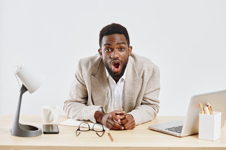Excited young man with dark skin sitting at office desk looking surprised and engaged, wearing beige...