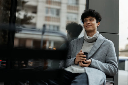 Young man sitting on bench in front of building with hands in pockets and cup of coffee in hand