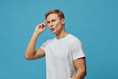 Confused man making gesture with fingers near temple in casual white t-shirt, expressing uncertainty or doubt, isolated on blue background. People concept with focus on emotion and body language.の写真素材