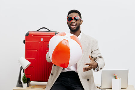 Vibrant young man with dark skin wearing stylish sunglasses and light blazer, holding a colorful beach ball, standing in front of a travel suitcase and desk, cheerful vibe. People lifestyle concept, travel preparation.の写真素材