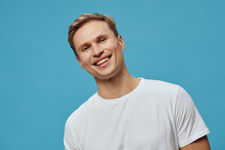 Smiling young caucasian man wear white t-shirt look camera in front solid blue background happy...