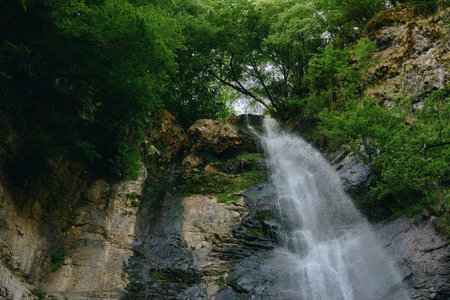 Beautiful natural waterfall cascading down rocky cliffs surrounded by lush green trees, creating a...
