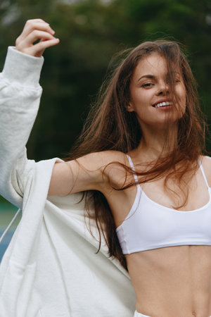 Young woman in a white sports bra and oversized cardigan enjoying a breezy day, exuding happiness and confidence in a nature backgroundの写真素材