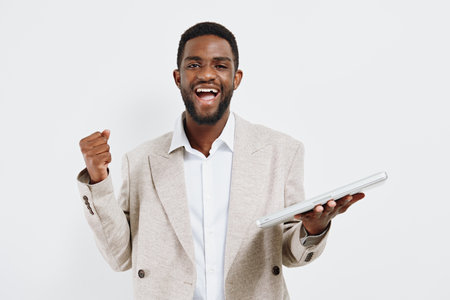 Smiling man with short curly black hair and beard, wearing a light beige checked blazer and white shirt, expressing excitement and success, holding a laptop in one hand, raising his fist, shot against a plain white background, studio portrait. People lifestyle conceptの写真素材
