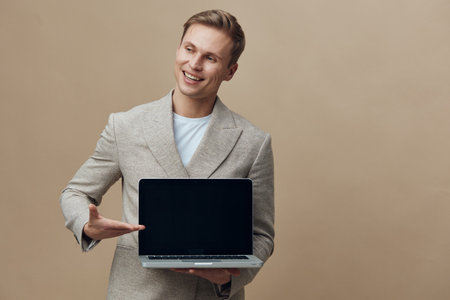 Confident young Caucasian man in smart casual blazer holding open laptop computer and pointing at screen isolated on beige background studio portrait. Business technology conceptの写真素材