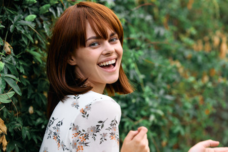 Vibrant woman with red hair smiling in front of lush green bush on sunny day