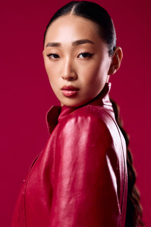 Fashion asian brunette in red leather jacket against pink background, with hair in ponytailの写真素材