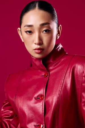 Fashionable asian brunette woman in a red leather jacket posing with hands on hips and head tilted against a pink backgroundの写真素材
