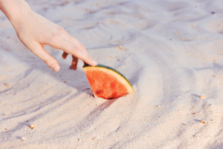 Hand reaching for a slice of watermelon on sandy beach backgroundの写真素材