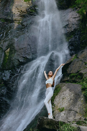 smiling woman in white activewear stretching arms near large waterfall surrounded by lush green rocks and mist, showcasing outdoor fitness and freedom.の写真素材