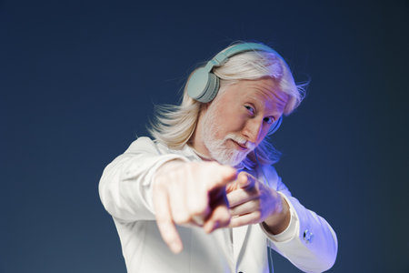 Elderly man with long gray hair and headphones, confidently pointing at the viewer against a dark blue background, conveying energy and modernity through stylish attireの写真素材