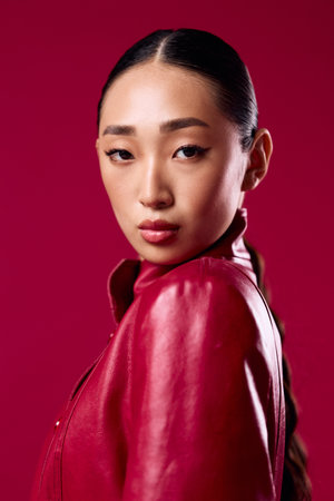 Fashion asian brunette in red leather jacket posing against pink background with hair pulled back in a ponytailの写真素材