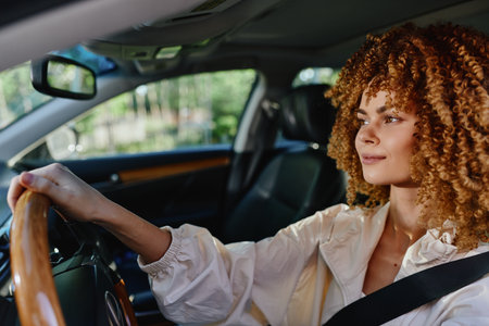 Young woman driving car with curly hair, wearing seatbelt and light blouse, focused and calm, natural light inside modern vehicle interior.の写真素材