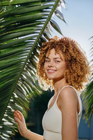 smiling woman with curly hair enjoying sunlight outdoors near tropical palm leaves, wearing white top and expressing happiness on a bright sunny dayの写真素材