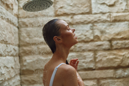 woman showering outdoors with closed eyes under rain showerhead, wet hair, stone wall background, natural light, serene mood, summer wellness momentの写真素材
