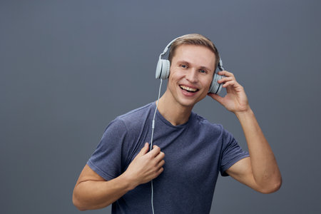 Happy young man with short hair and casual blue T-shirt wearing large over-ear headphones enjoying...