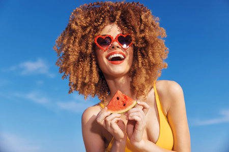 Cheerful woman with curly hair holding a slice of watermelon in sunglasses against a bright blue sky