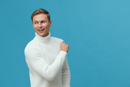 Delightful smiling man with light brown hair wearing cozy white knit sweater, expressing happiness and friendliness, isolated on vibrant blue background, studio portrait, casual lifestyle concept.の写真素材