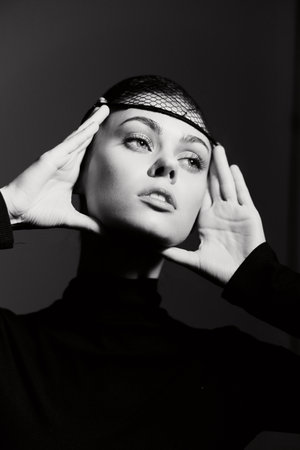 Elegant young woman in black, wearing a fishnet headpiece, posing with a contemplative expression against a dark background. Fashion and beauty concept.の写真素材