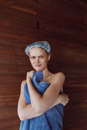 Young woman wrapped in blue towel with shower cap on, standing against wooden wall, smiling softly in warm indoor bath setting, natural beauty and relaxation atmosphere.の写真素材