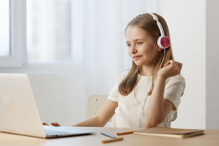 Young girl with headphones enjoying music while using a laptop in a bright, minimalist workspace,...