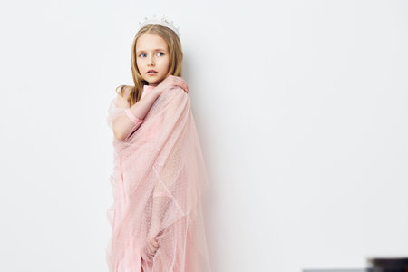 Young girl in a soft pink dress with a crown against a white background, expressing innocence and joy, perfect for children s photography and concepts of childhoodの写真素材