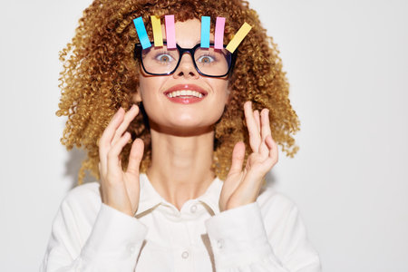 Cheerful woman with curly hair and colorful clothespins on glasses, smiling against a light grey background, showcasing a playful and quirky styleの写真素材