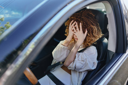Stressed woman covering face with hands inside car wearing seatbelt with curly hair and white blouse, expressing frustration or anxiety during daytime drive.の写真素材