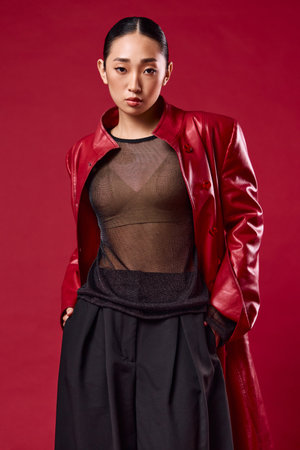Fashion asian woman in red leather jacket and black pants posing with eyes closed against red backgroundの写真素材