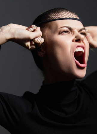 Angry woman expressing intense emotions, wearing a black top and fishnet headpiece against a dark background. Concept of frustration and emotional release.の写真素材