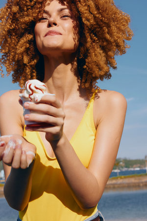 Young woman with curly hair enjoying ice cream on a sunny day by the waterの写真素材