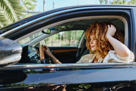 Curly woman driving a black car, smiling happily with casual outfit and natural sunlight, relaxing outdoors on a sunny day during spring season.の写真素材
