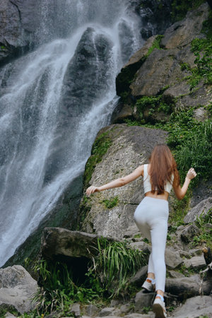 woman hiking near waterfall in athletic wear climbs rocky trail surrounded by greenery and flowing water in outdoor adventure sceneの写真素材