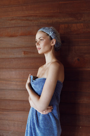young woman wrapped in blue towel with shower cap standing contemplative against wooden wall in spa...